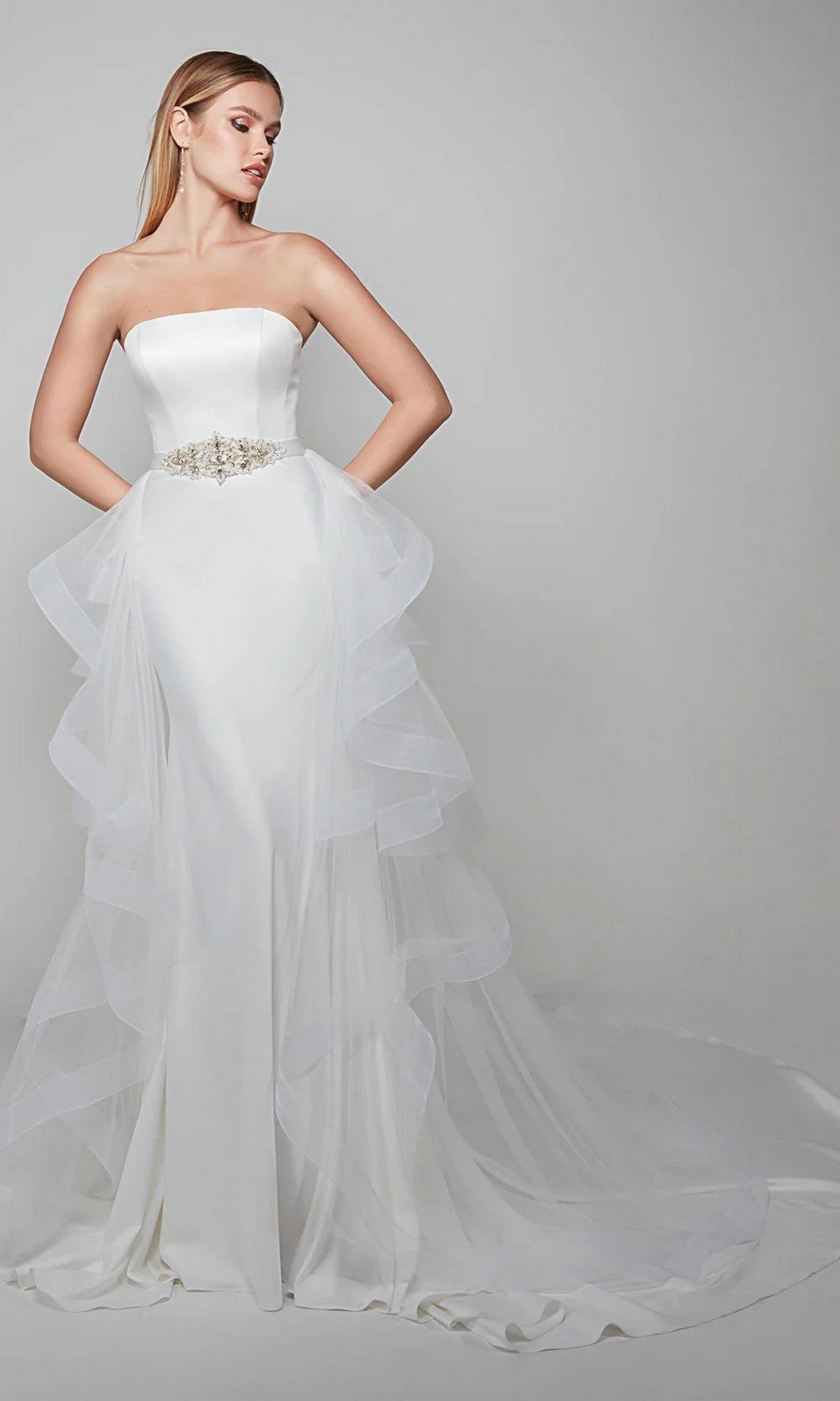A woman wears the Alyce 7025 long white dress, a strapless formal gown with a fitted bodice, waist belt, and layered tulle overskirt with a long train, standing against a plain backdrop.